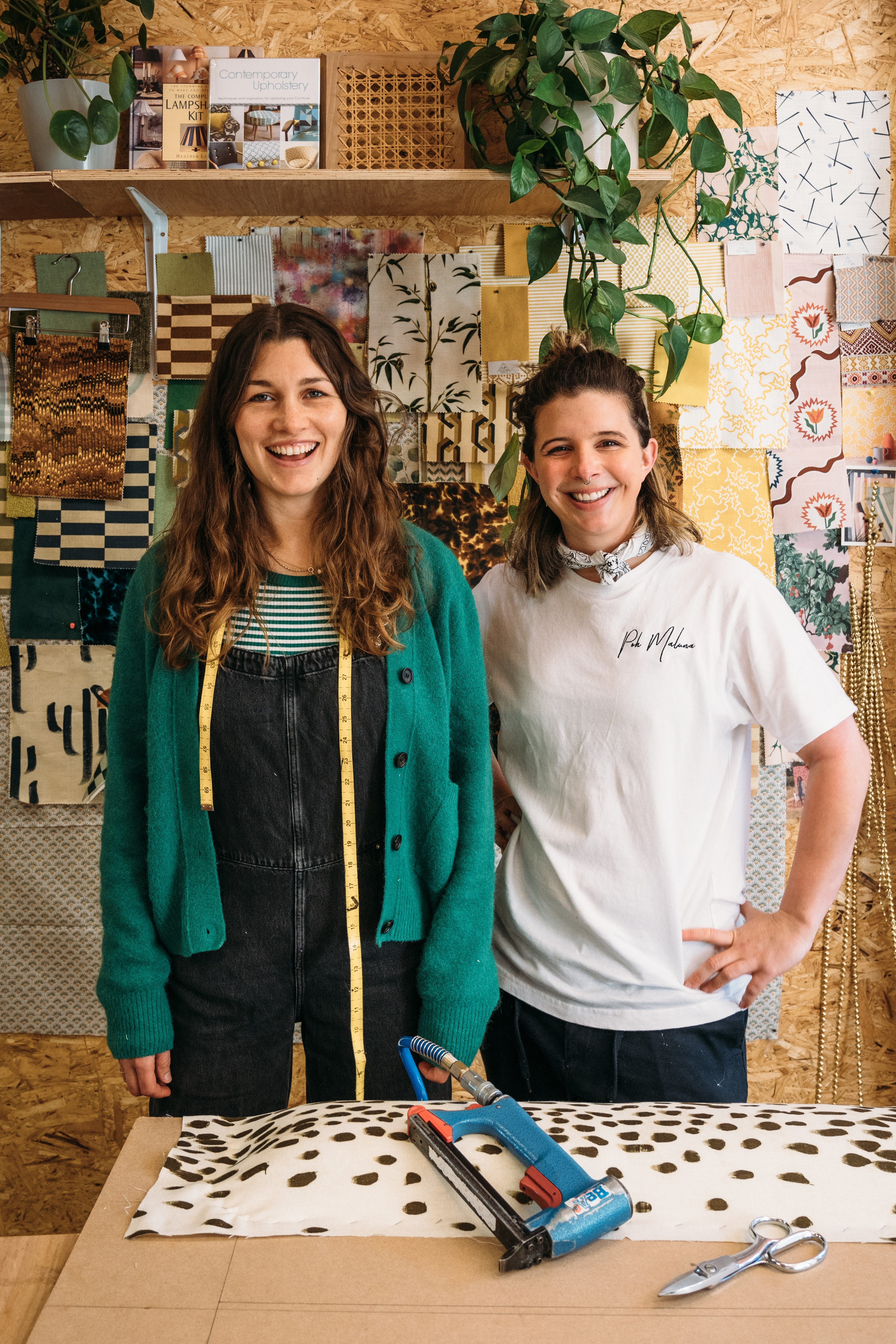 Jo and Jess smiling in front of their workbench with fabric and upholstery tools in front of them.