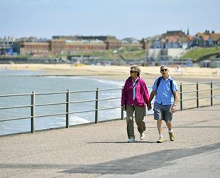Man and lady walking along promenade holding hands. Man in blue shirt and grey shorts. Lady wearing a pink fleece and beige trousers. Blurred background of railings, beach, sea and buildings