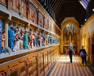 Man and lady looking at wooden panelling with coloured carvings showing depicting bible scenes. Underneath brass plaques. At end of small alter