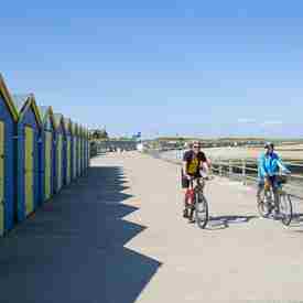 Couple cycling along prom at Minnis Bay in front of blue and yellow beach huts with beach on the right
