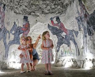 Two little girls and mum crouched down in white chalk caves pointing at artwork on the walls. Behind artwork of men on horses taking part in a hunt