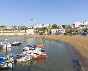 Looking from harbour across empty sandy beach with small boats and yachts in the harbour. Row of colourful beach huts at bottom of cliffs and promenade. Bright blue sky 