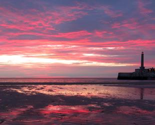 Pink sunset sky above Margate stone harbour arm. Tide out with large area of sand in front of arm and boast in the harbour. Lights on outside the buildings on the arm