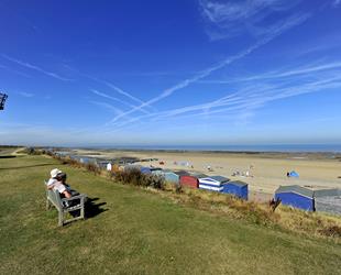 Couple sat on wooden bench on the grass above the beach, looking out across the beach and sea. Bright blue sky