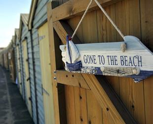 Wooden 'Gone to the Beach' arrow sign with blue and white stripes hanging on beach hut door with other beach huts in the background