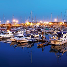 Twilight Ramsgate Royal Harbour with vivid blue sky and sea, golden lights shining from boats, buildings, arches and road. White railings around harbour edge. Variety of boats and yachts in harbour and traffic travelling along road  