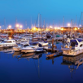 Twilight Ramsgate Royal Harbour with vivid blue sky and sea, golden lights shining from boats, buildings, arches and road. White railings around harbour edge. Variety of boats and yachts in harbour and traffic travelling along road
