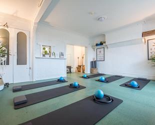 White room with turquoise floor. Grey yoga mats, with steps, rings and small blue balls on them laid out. Posters of yoga moves on the wall and green tall plants in the room corners. 