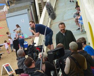 Two ladies and group of children on the beach at the side of a beach hut, Man eating ice cream and holding a windmill leant on white wooden railing. Actor talking to camera with group of film crew behind the camera