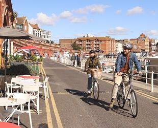 Couple cycling along Ramsgate seafront with people sat at tables. Harbour with boats on the right handside