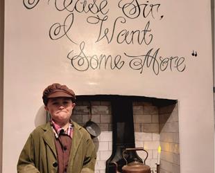 Boy in Dickens costume stood in front of oven with 'Please Sir I want some More' on wall behind