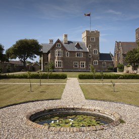 Gravel path around circular pond leading through lawn towards brick gothic house with flag flying from tower turret.  Young trees and box hedge across grass. Bright blue sky.