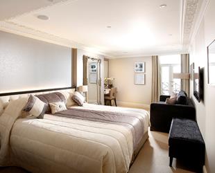 King size bed with white coverings and bedside unit with lamp. Large black box at end of bed next to large black sofa. Large wall mirror opposite sofa with white desk to side. Long large window in right corner