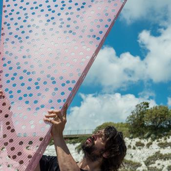 Man stood holding pink metal triangle with holes in, part of sculpture. Behind chalk cliffs, trees and bright blue sky 