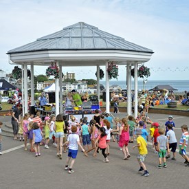 Children dancing in front of white bandstand with dj playing music. Variety of gazebos/stalls either side of bandstand selling/promoting things.  Light blue/grey sky