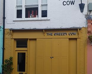 Dark yellow painted door and window shutters with white writing 'The Greedy Cow'. Above white painted wall with black writing of The Greedy Cow and black old style street lamp