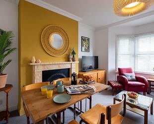 Bright yellow chimney breast behind wooden table and chairs. TV on unit on right behind red chair and table by window