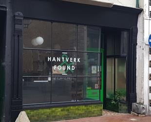Black surround shop front with large clear glass windows in grid format. White text in centre panel Hantverk & Found. Bright green entrance door