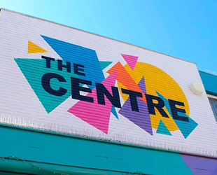 White background with brightly coloured triangles, circles and black text of The Centre on brick wall above shops