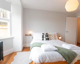 White room with large white bed in centre with green throw and cushions. Wooden floor with white rug next to bed and door on right.  Dark metal radiator under window on left side 