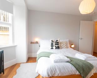 White room with large white bed in centre with green throw and cushions. Wooden floor with white rug next to bed and door on right. Dark metal radiator under window on left side