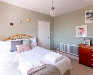 Pale grey walls with white door. Radiator and chest of drawers below pictures on right side. Double bed in middle with white coverings and 3 pastel coloured cushions. Glass pineapple room light