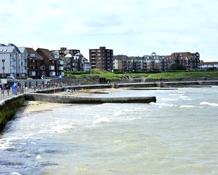 Looking across the beach with the tide up covering most of the beach. People walking along the promenade. The beach is located down in front of grass and garden banks in front of a road and properties 